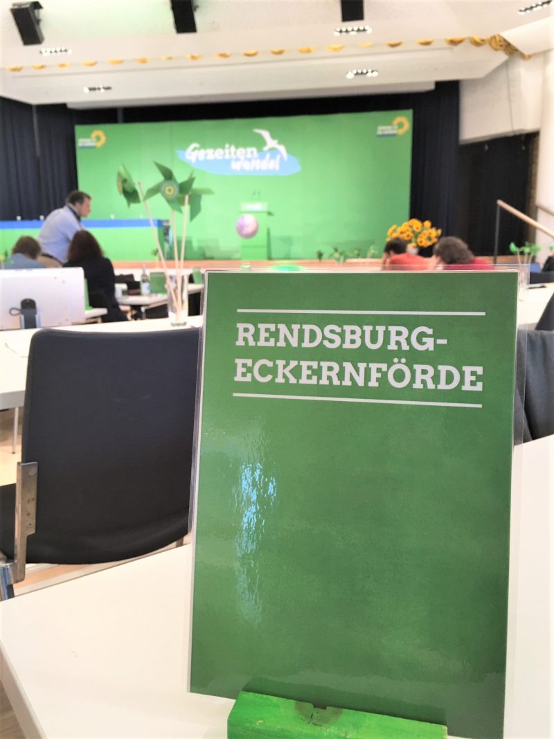 Das Bild zeigt im Vordergrund eine grüne Tischkarte, auf der Rendsburg-Eckernförde geschrieben steht. Der Hintergrund ist unscharf. Man erkennt einen Saal mit Tischen und Stühlen, im Hintergrund ist eine Bühne. Darauf ein Rednerpult. Die Bühne ist leer. Auf einer grünen Wand steht das Motto des Parteitages: Gezeitenwandel.