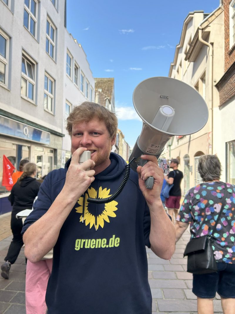 Dennis aus dem Ortsvorstand des Ortsverbandes Region Rendsburg hält ein Megafon und trägt einen Pull mit der Aufschrift "gruene.de", im Hintergrund sind weitere Demo-Teilnehmende zu sehen